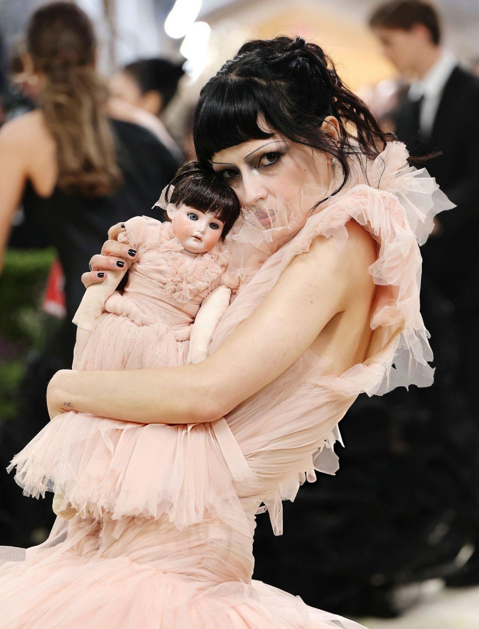 Woman in a pale pink tulle dress cradles a porcelain doll wearing a matching pink outfit at a formal event.