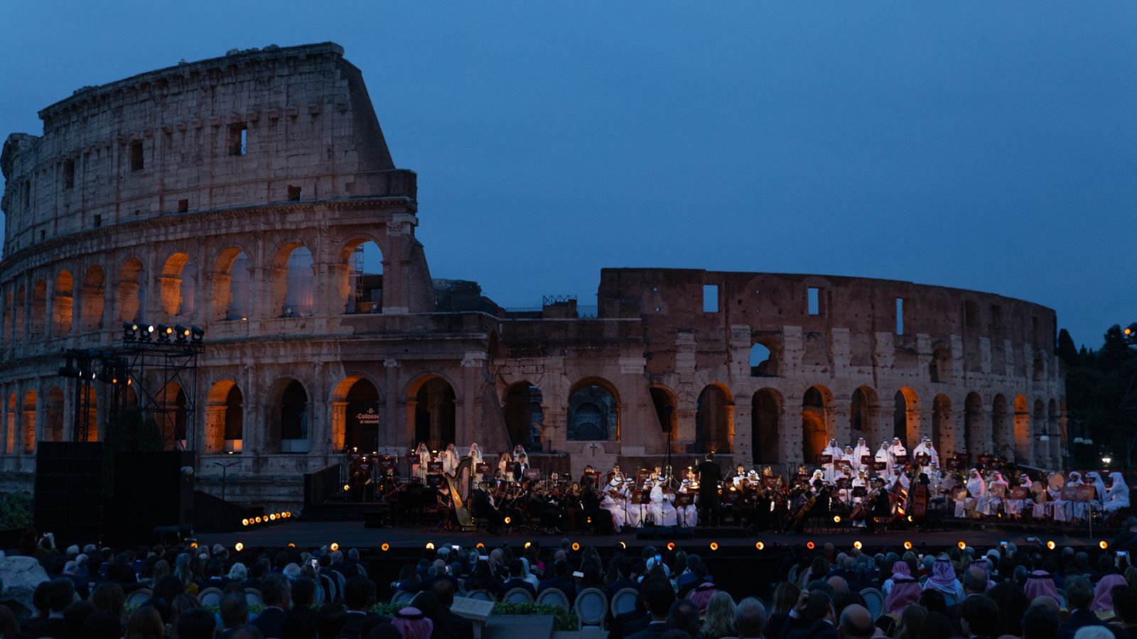 Orchestra performing on stage in front of the Colosseum at dusk, audience seated in foreground.