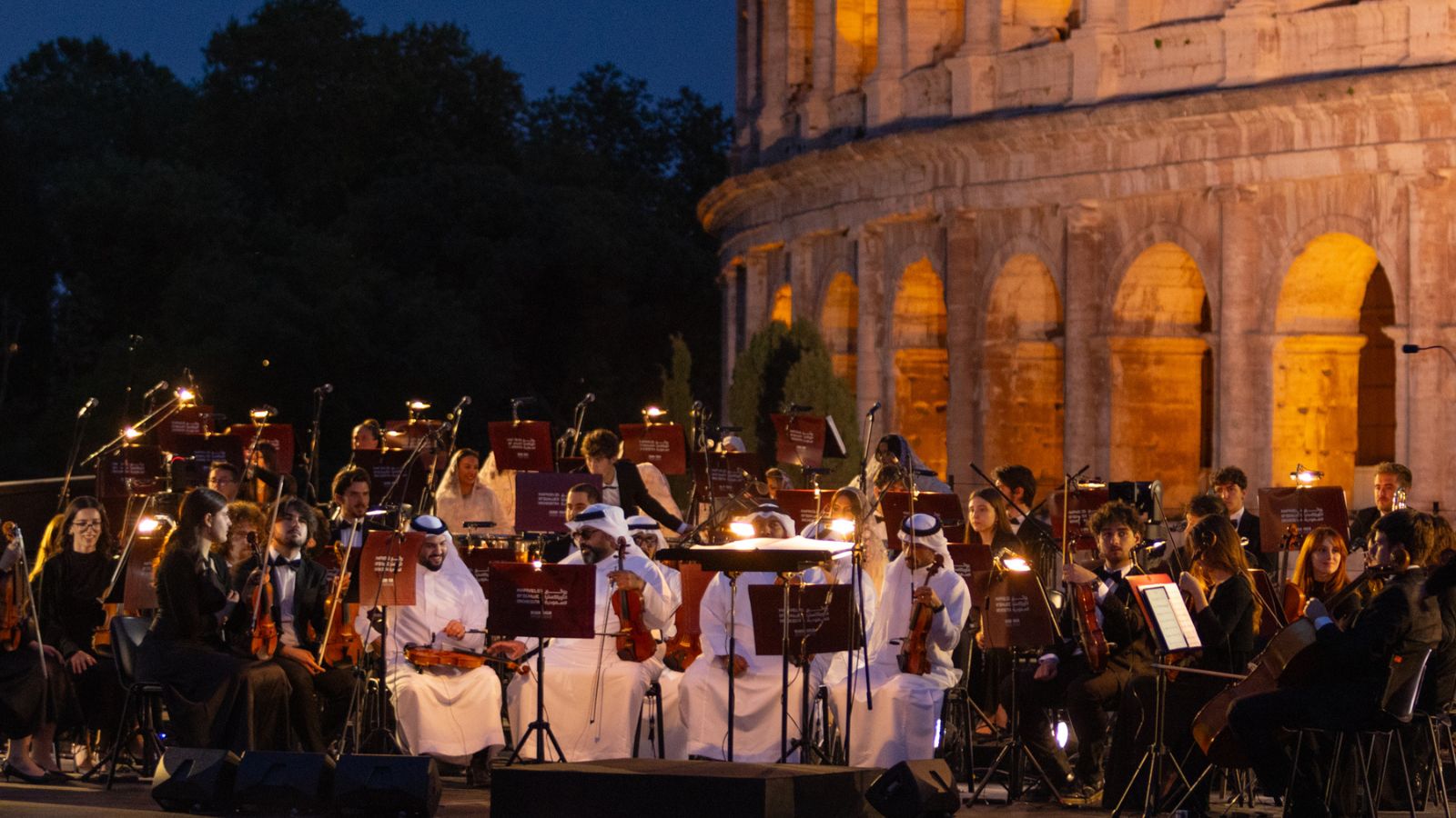 Outdoor orchestra performance at night beside a lit ancient stone amphitheater; musicians with violins, cellos, and sheet music on stands.