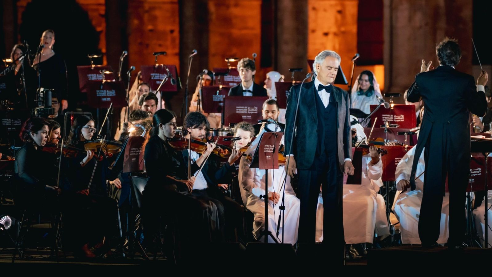 Conductor in a tuxedo directs a seated orchestra under warm stage lighting during a concert.