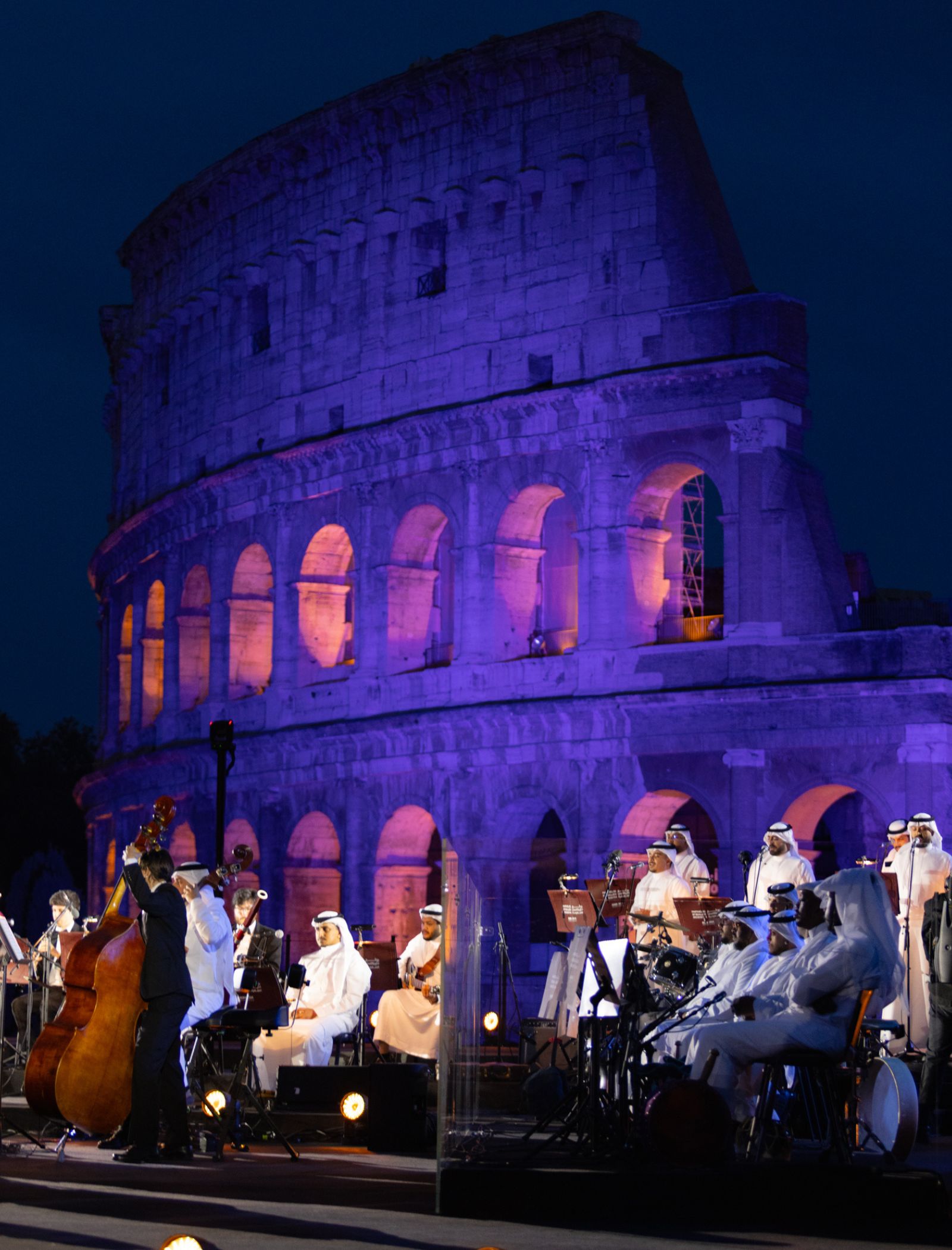 Night view of the Colosseum lit in purple and orange with a large orchestra performing on stage in front of it men in white robes playing instruments.