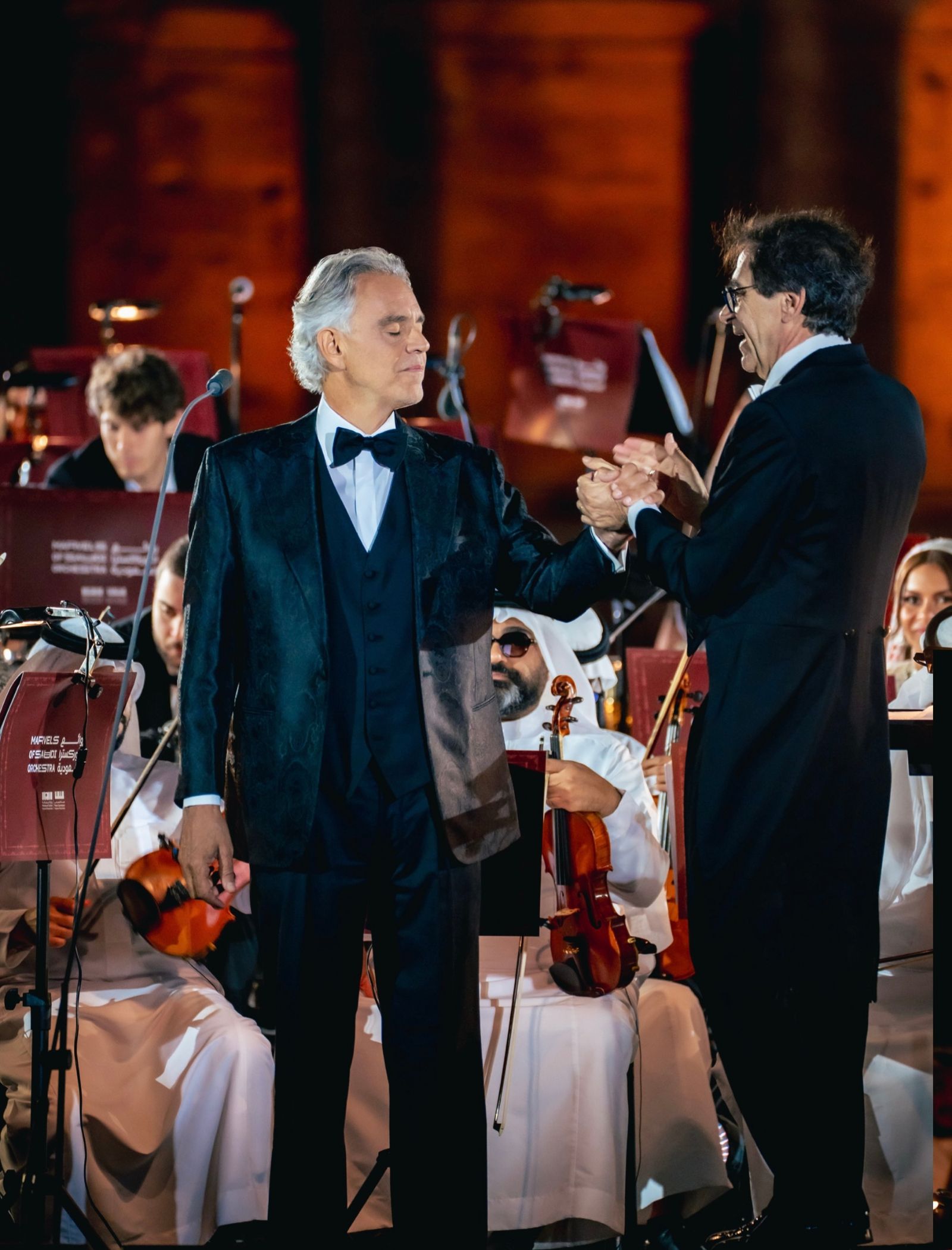 Two men in tuxedos shake hands on a concert stage, with musicians performing in the background.