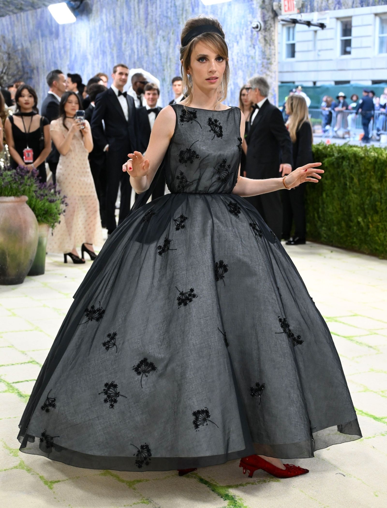Woman in a voluminous gray ball gown with black floral embroidery posing on a red carpet, with guests and photographers in the background.