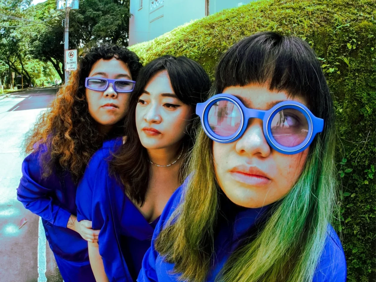 Three friends pose for a close selfie outdoors, wearing blue outfits and oversized blue glasses, with greenery in the background.