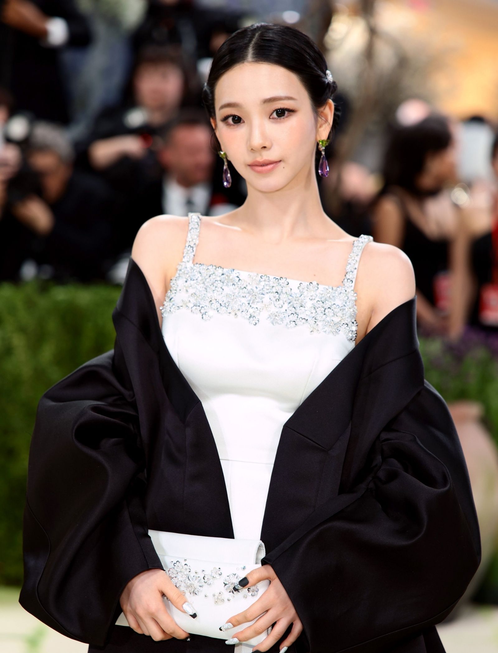 Woman on a red carpet in a silver-embellished white gown with a black wrap, holding a shimmering clutch.