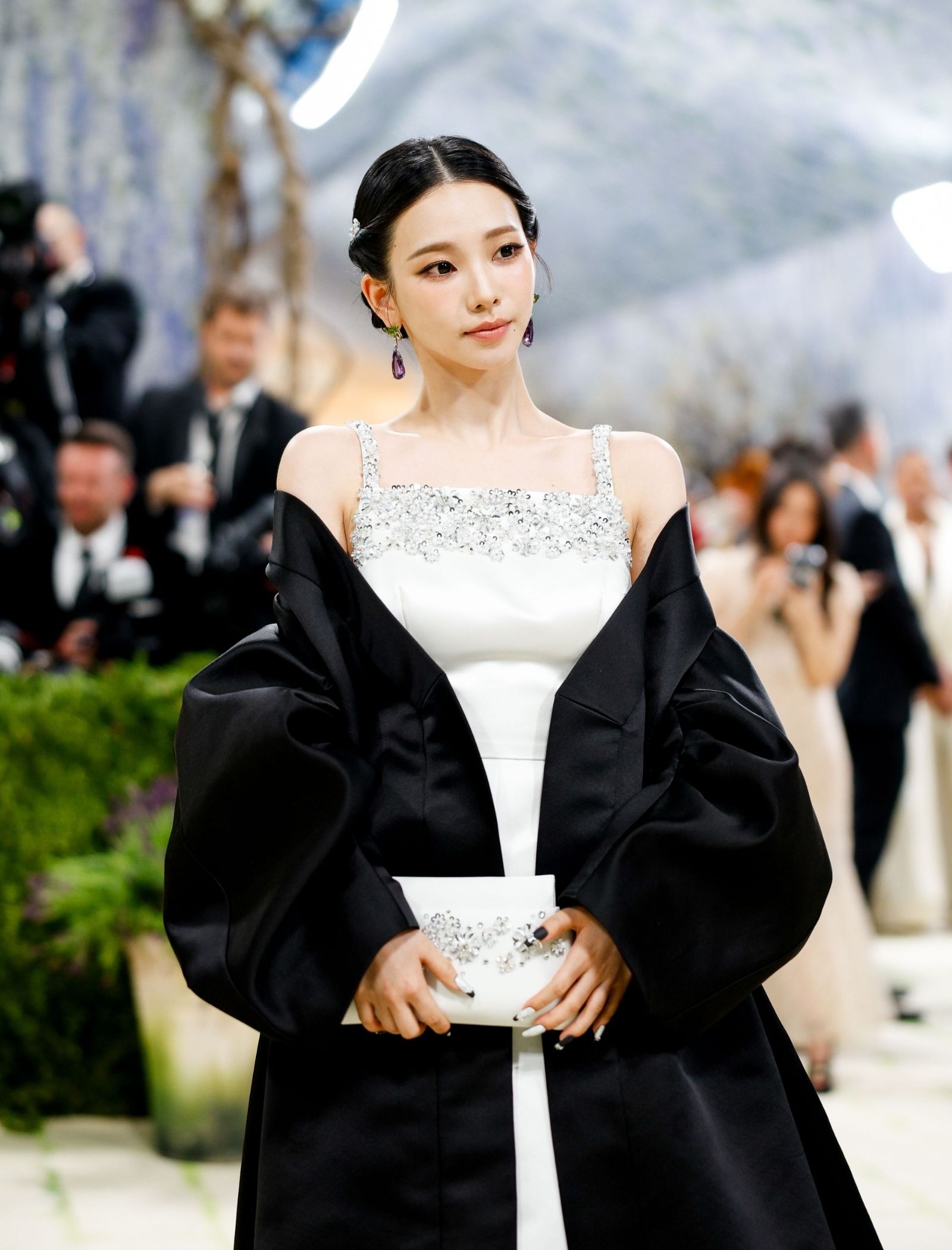 Elegant woman on a red carpet wearing a white gown with silver beading and a black shawl, posing for cameras.