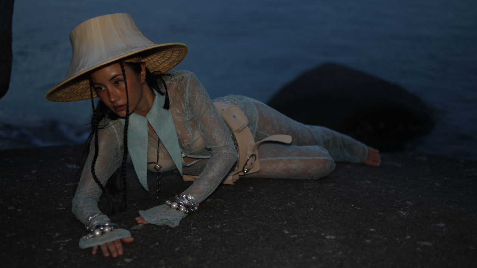 Woman in a wide-brim straw hat and mint lace outfit lying on a rocky shoreline by dark water at dusk/nighthood.