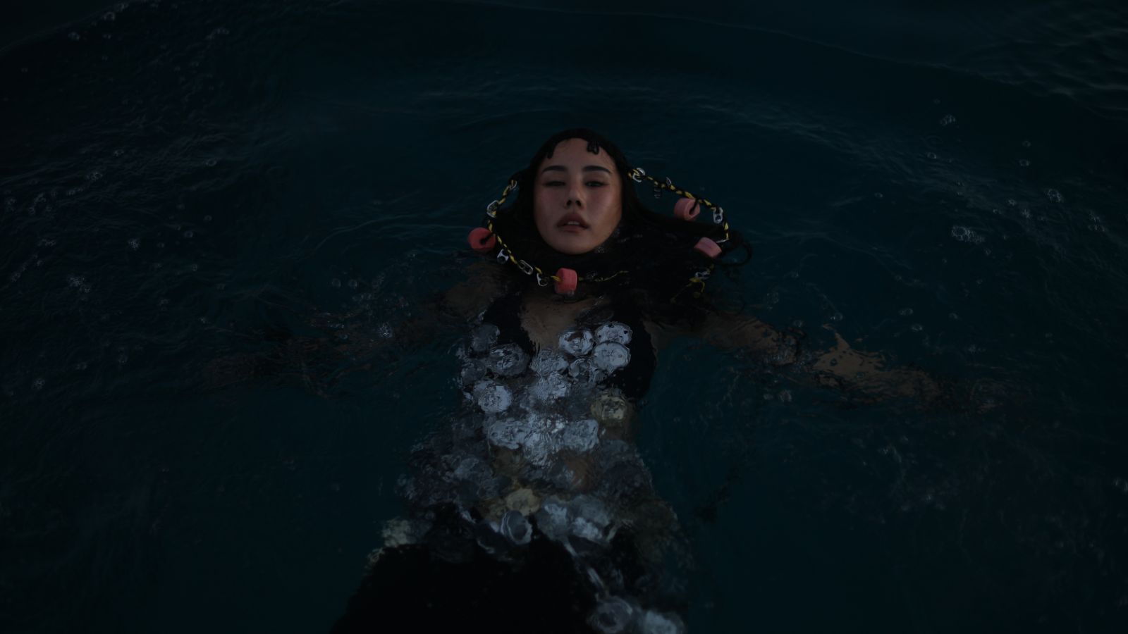 Person floating in dark blue water with head above surface, wearing a chain necklace with pink segments around the neck.