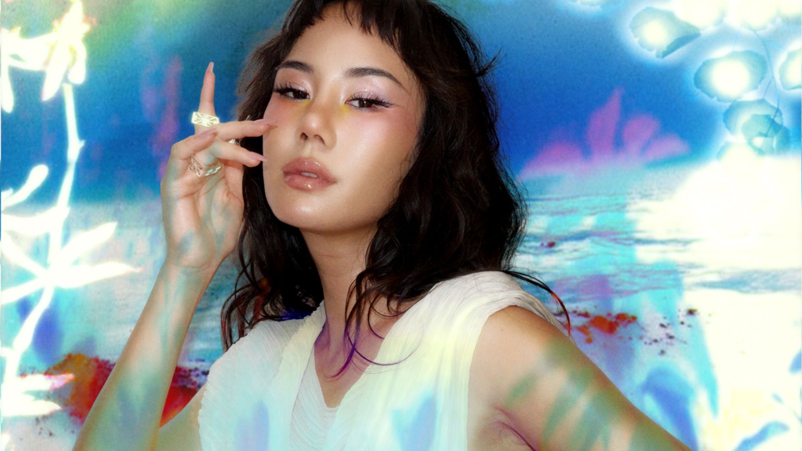 Portrait of a woman with dark wavy hair and glossy makeup, wearing a white sleeveless top, posing with her hand near her face against a vibrant abstract backdrop] ,