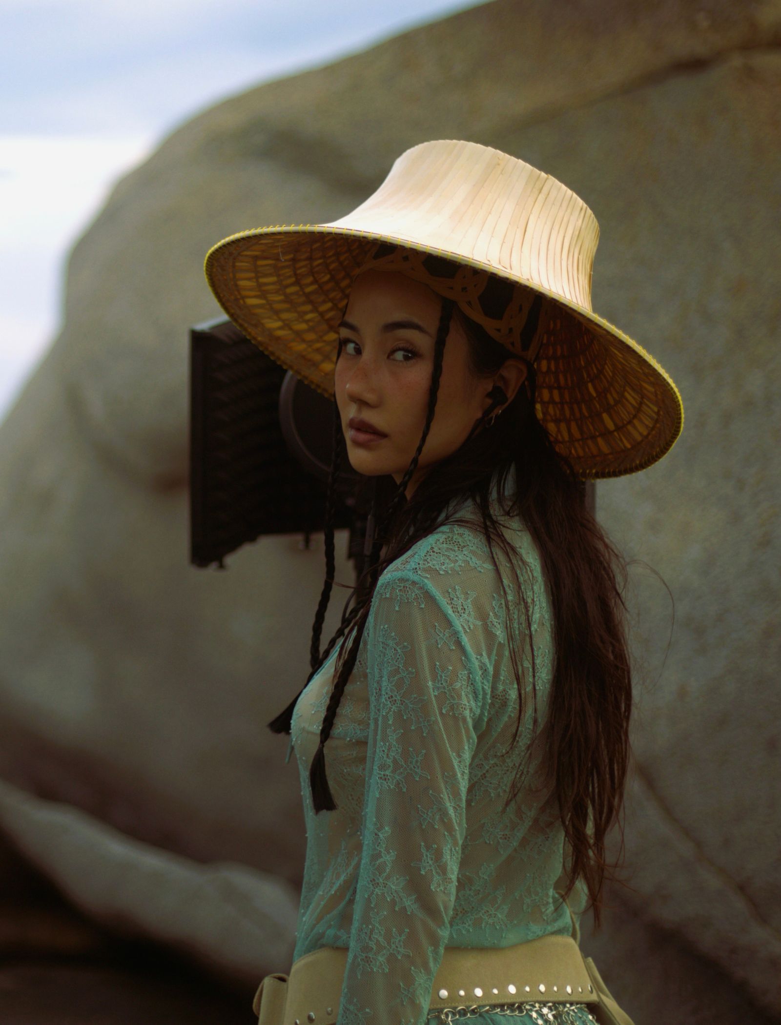 Woman wearing a large straw hat and a turquoise lace top, standing outdoors with rocky background.