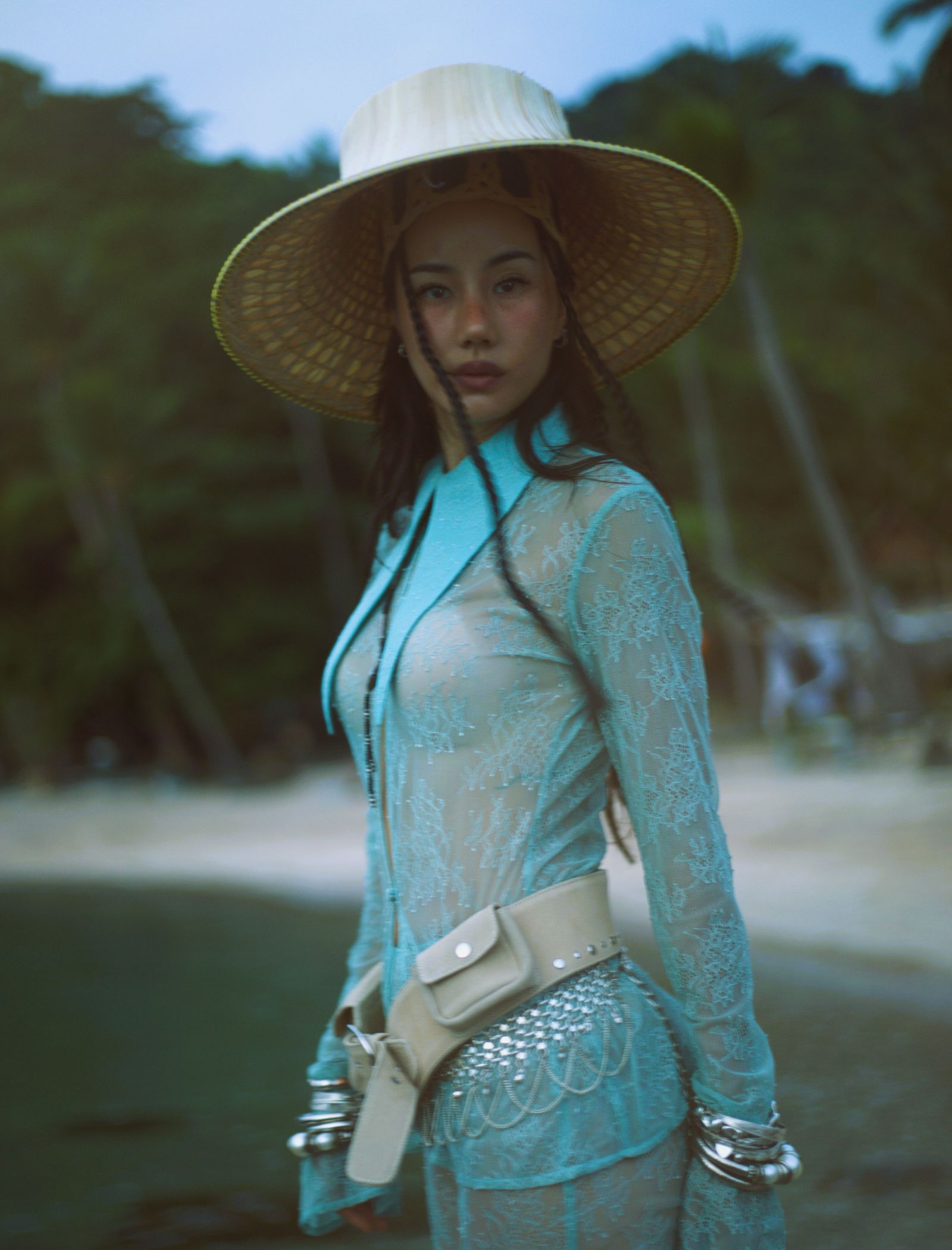 Woman in a turquoise lace dress with a large straw hat outdoors, facing slightly to the side.