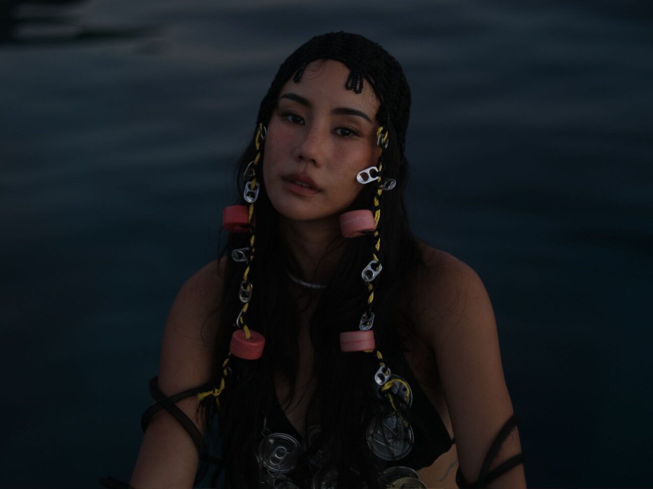 Portrait of a young woman with braided hair and colorful beads against a dark blue ocean backdrop at dusk.
