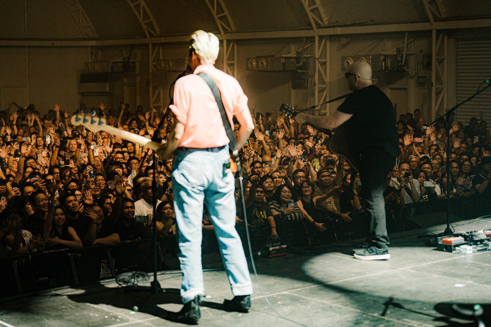 Two musicians perform on a crowded indoor stage as a large audience raises hands and smartphones in the foreground. One wears a pink shirt and white cap, playing guitar; the other in black and white sneakers stands to the side with a microphone and guitar equipment visible.