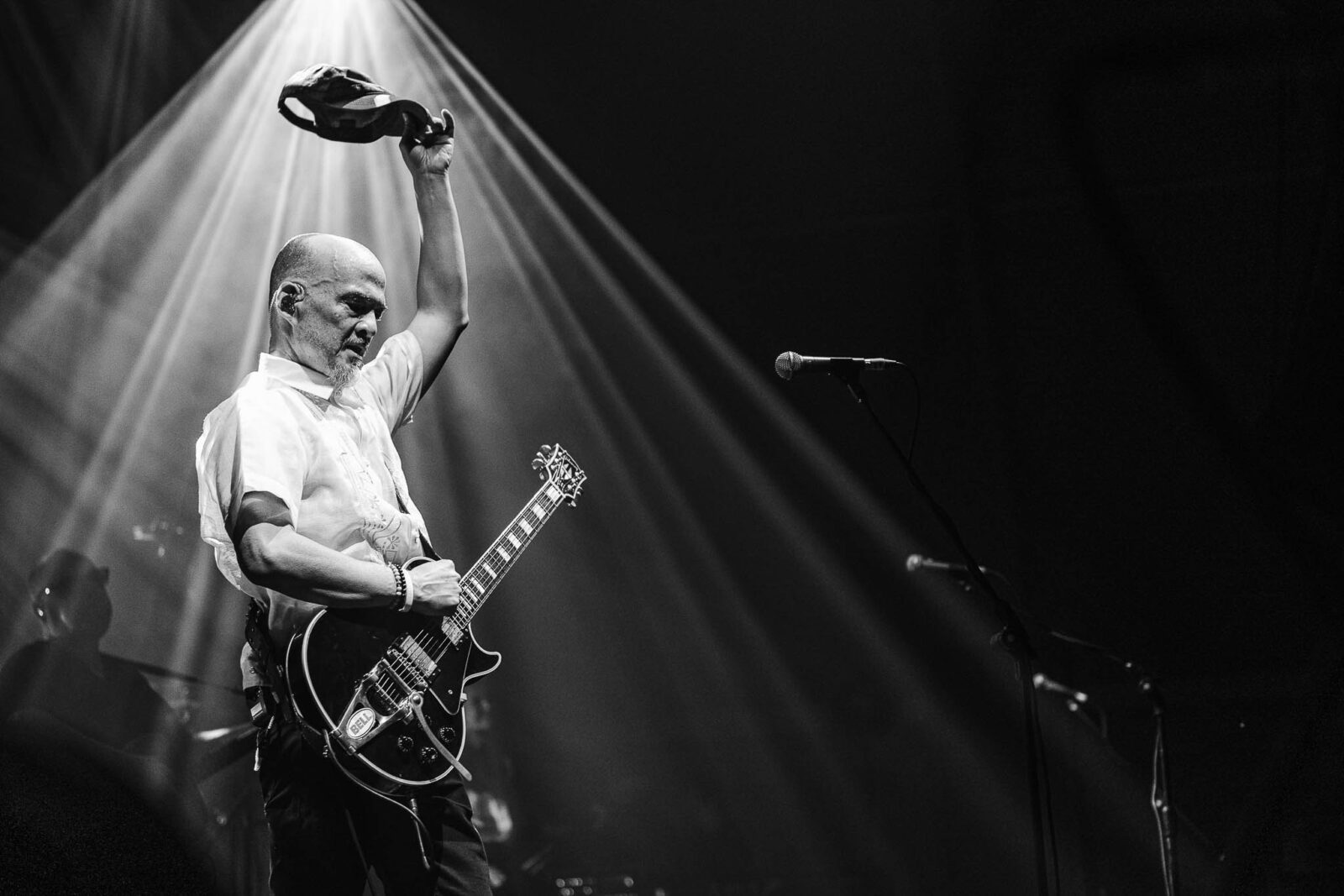 Guitarist on a lit stage, bald head, raising a hat while playing a black electric guitar under a spotlight.