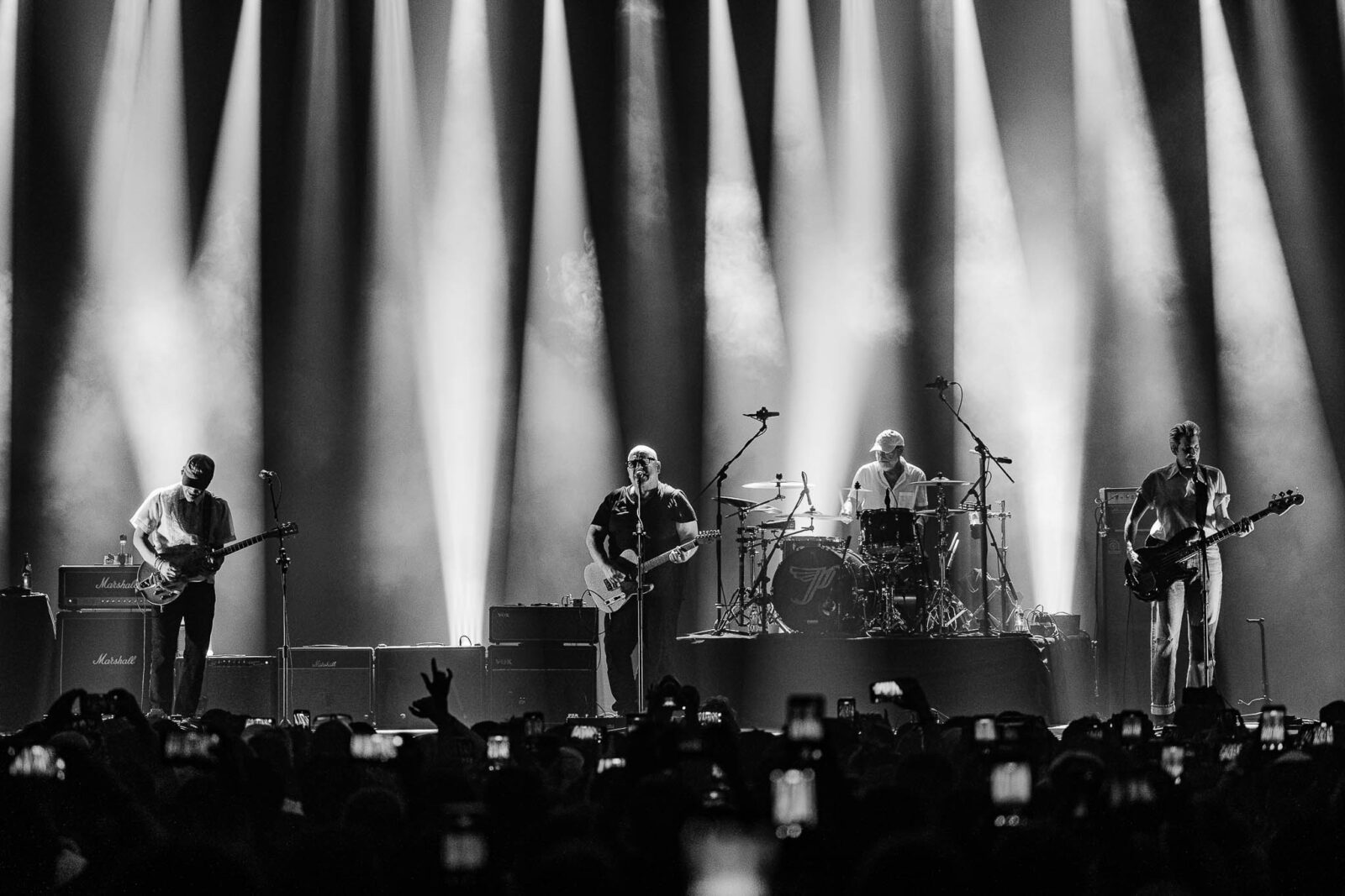 Black-and-white concert photo of a band performing on stage under dramatic vertical light beams, audience silhouettes in the foreground.