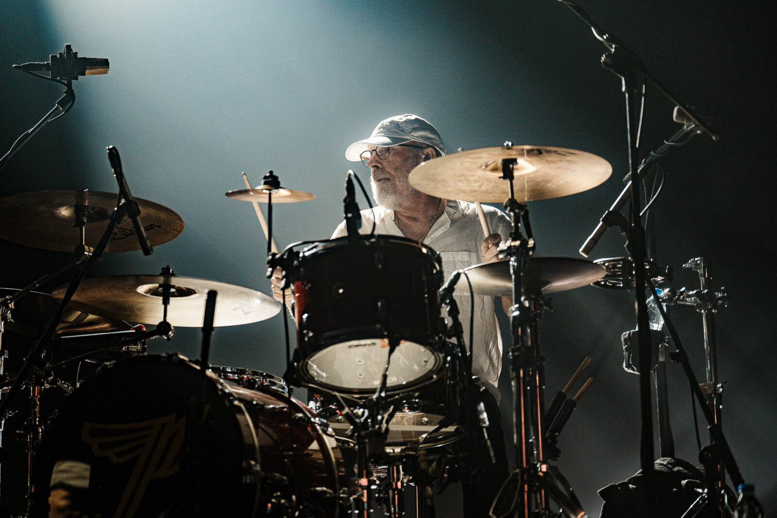 Drummer wearing a light cap and glasses playing a drum kit on stage with cymbals and microphones around him in moody lighting.