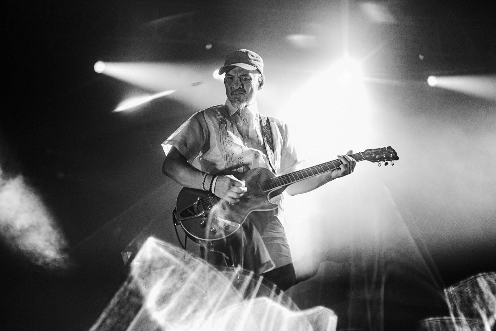 Male guitarist wearing a cap and light shirt performing on a dimly lit stage with bright backlights behind him.