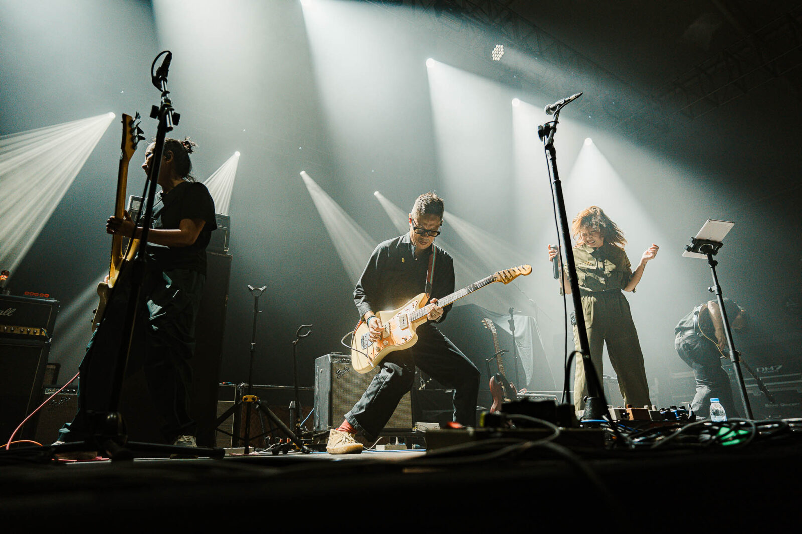 Live rock band on stage with guitarist center, bassist left, and female vocalist to the right, under dramatic white spotlights.