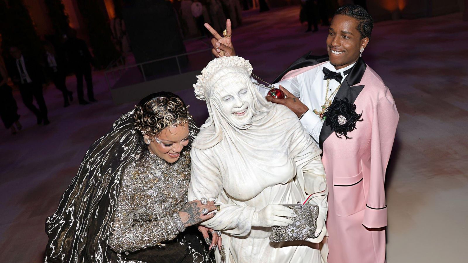 Three people pose together at a formal event: a person in a white sculpted gown with a headpiece, another in a sparkling beaded outfit, and a man in a pink suit making a peace sign.