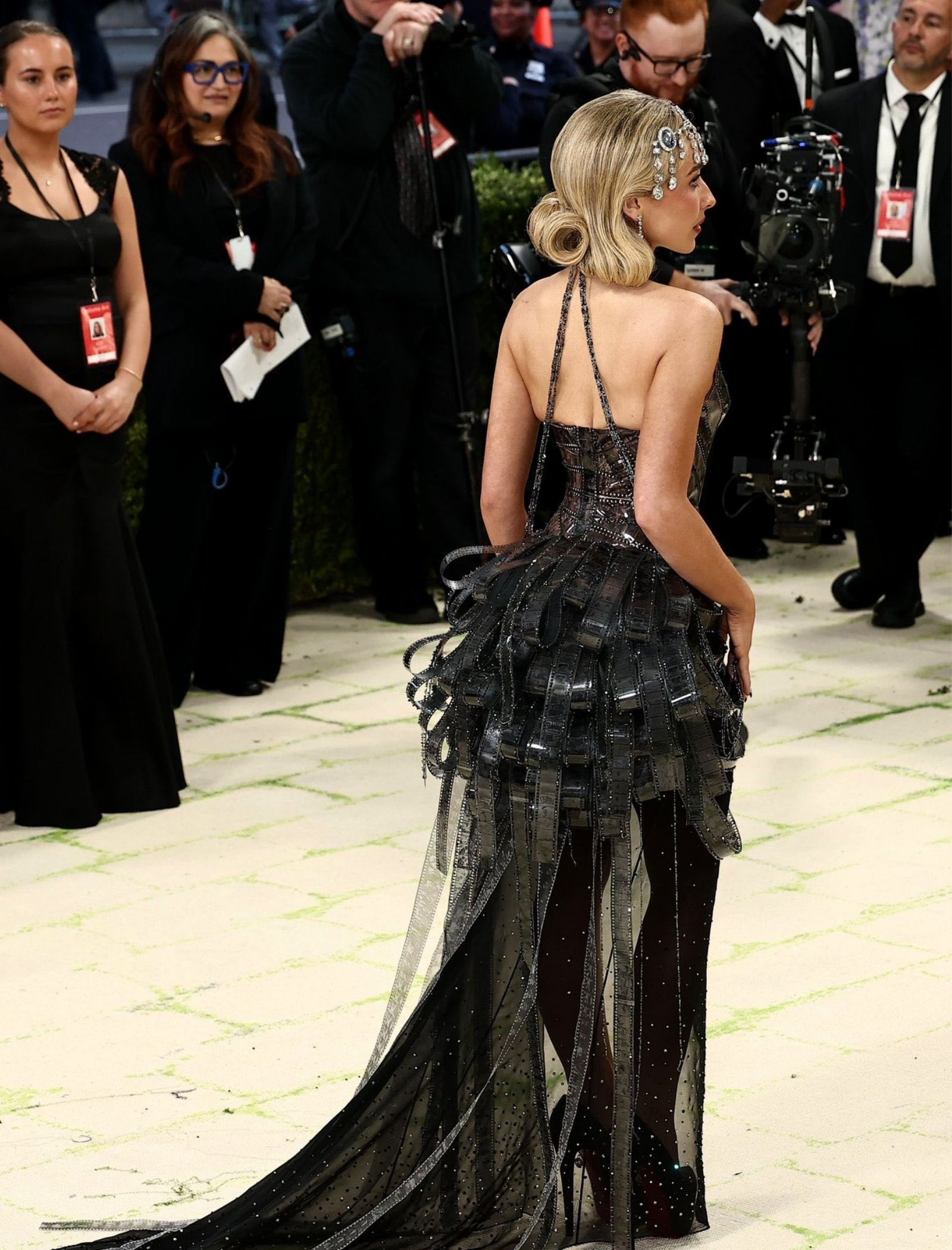 A woman in a black, sparkling gown with a sheer panel, her blonde hair styled in a retro bun, posing at a red carpet with photographers in the background.