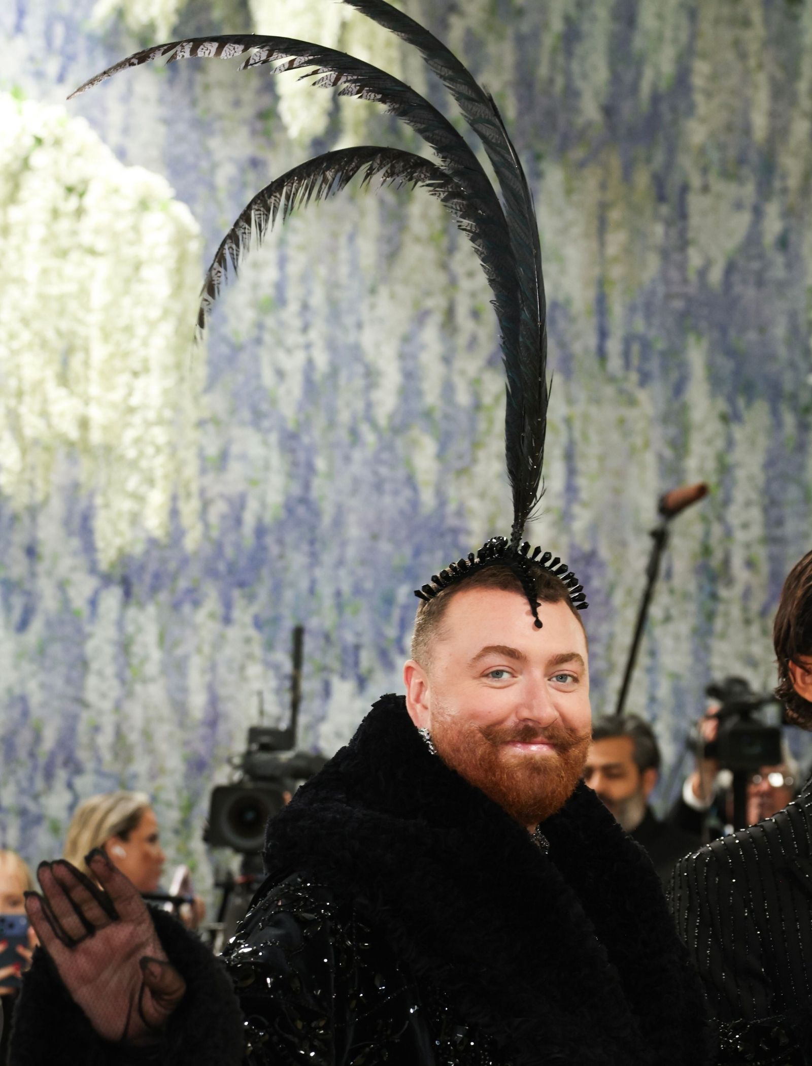 Man with a red beard wearing an elaborate black feather headpiece at a crowded event, smiling toward the camera with photographers in the background.
