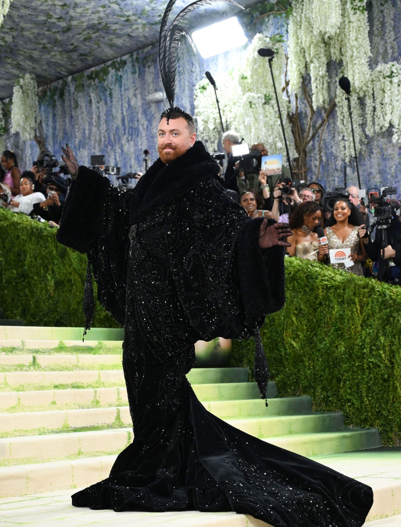 Man in an ornate black jeweled gown with a tall feather headpiece stands on a green-carpeted staircase, posing for photographers among reporters and cameras.