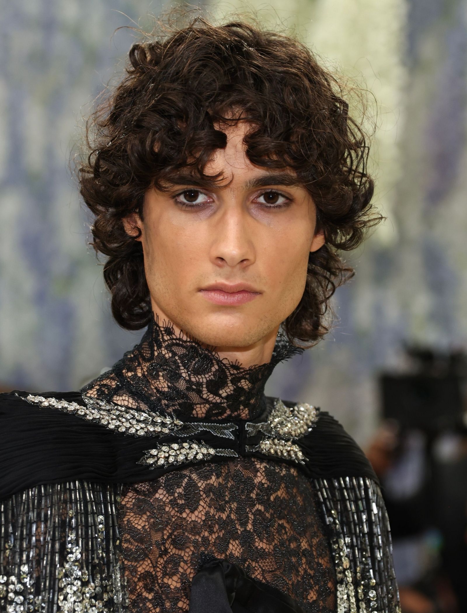 Close-up of a fashion model with dark, curly hair wearing a black lace high-neck top embellished with silver sequins on a runway backdrop.