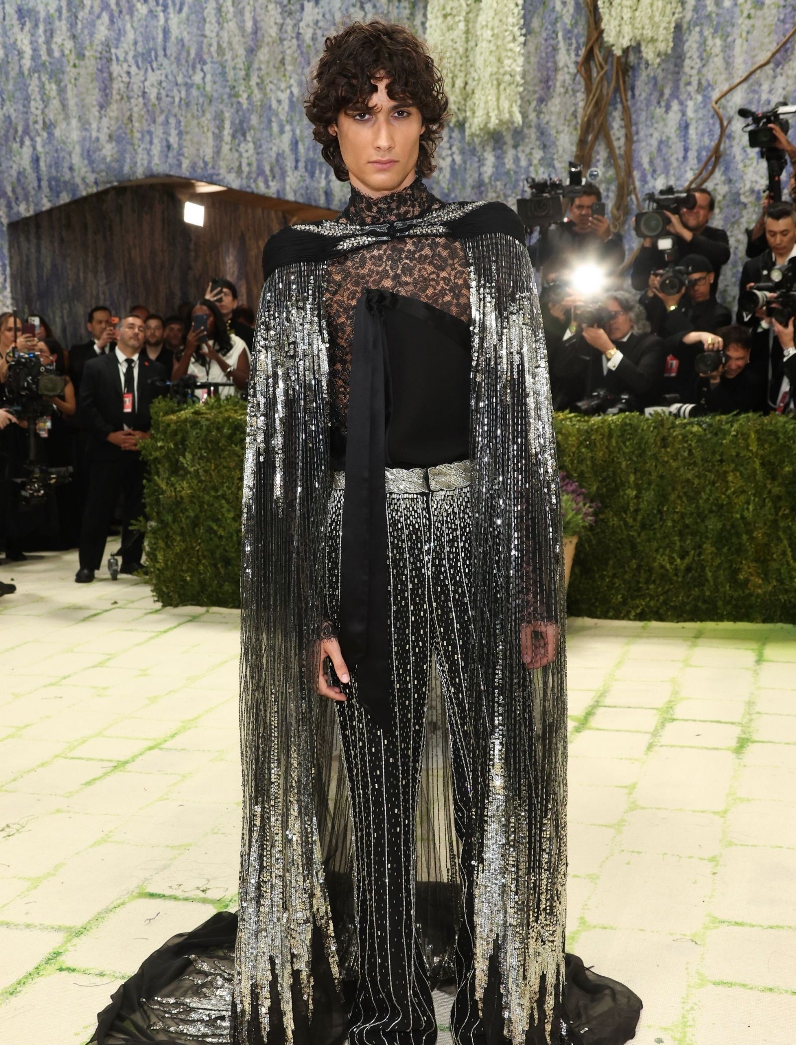 Model in black lace and silver fringe cape posing on a runway with photographers in the background