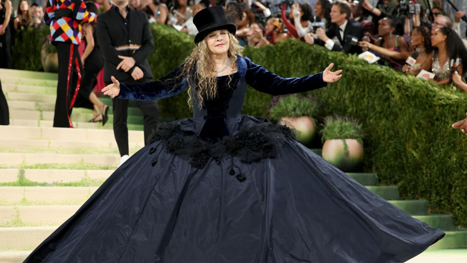 Woman in a dramatic navy ball gown with outstretched arms on a red carpet steps, wearing a top hat at a formal event.