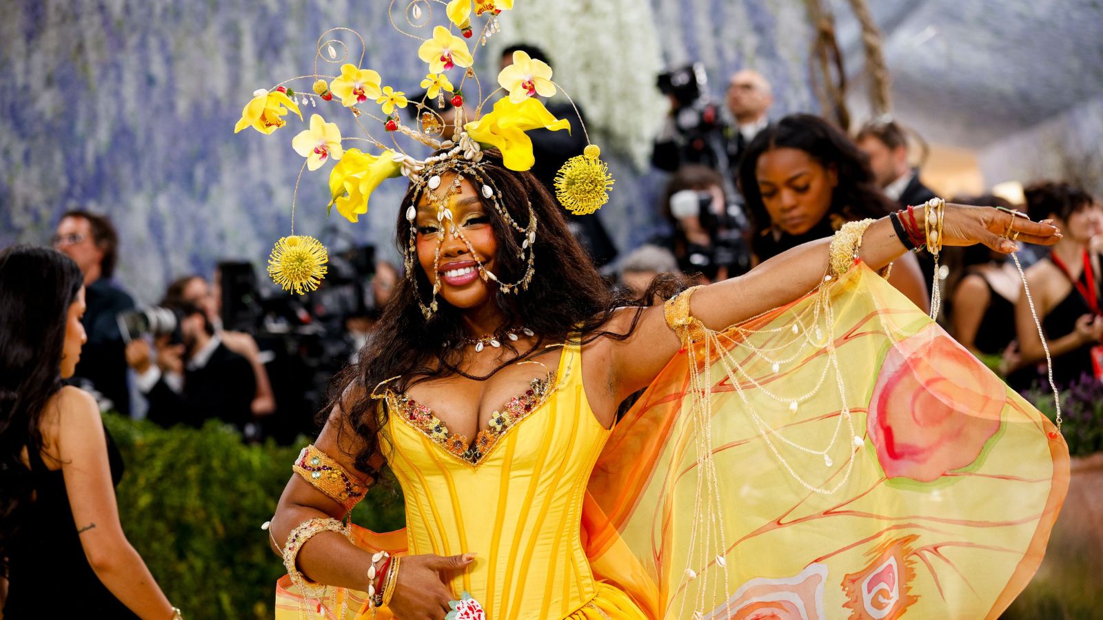 Woman in a bright yellow gown dancing on a red carpet with an elaborate yellow floral headpiece and flowing cape, photographers in the background.]}, {