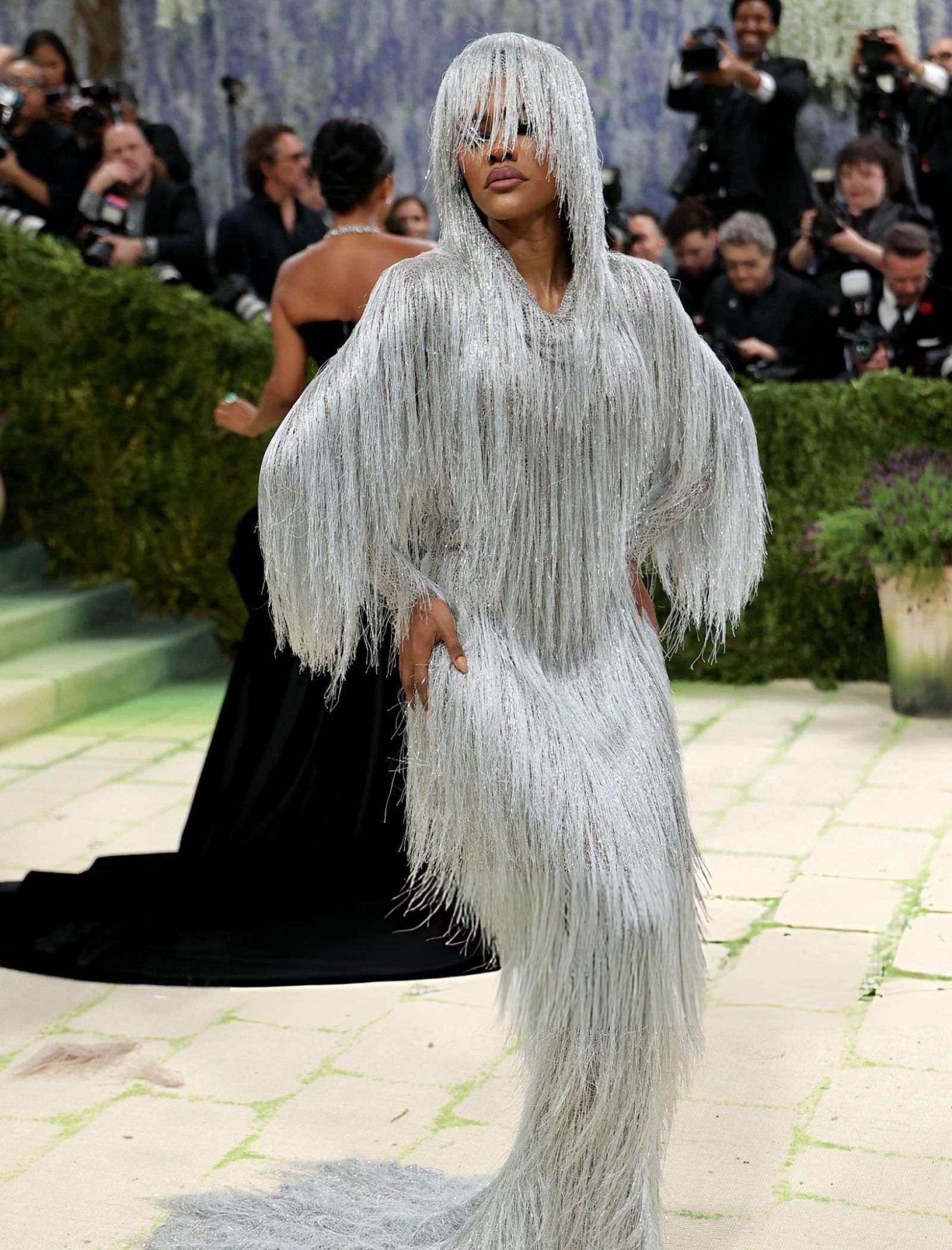 Person wearing a floor-length silver fringe gown with a hood, posing on a runway as photographers look on in the background.