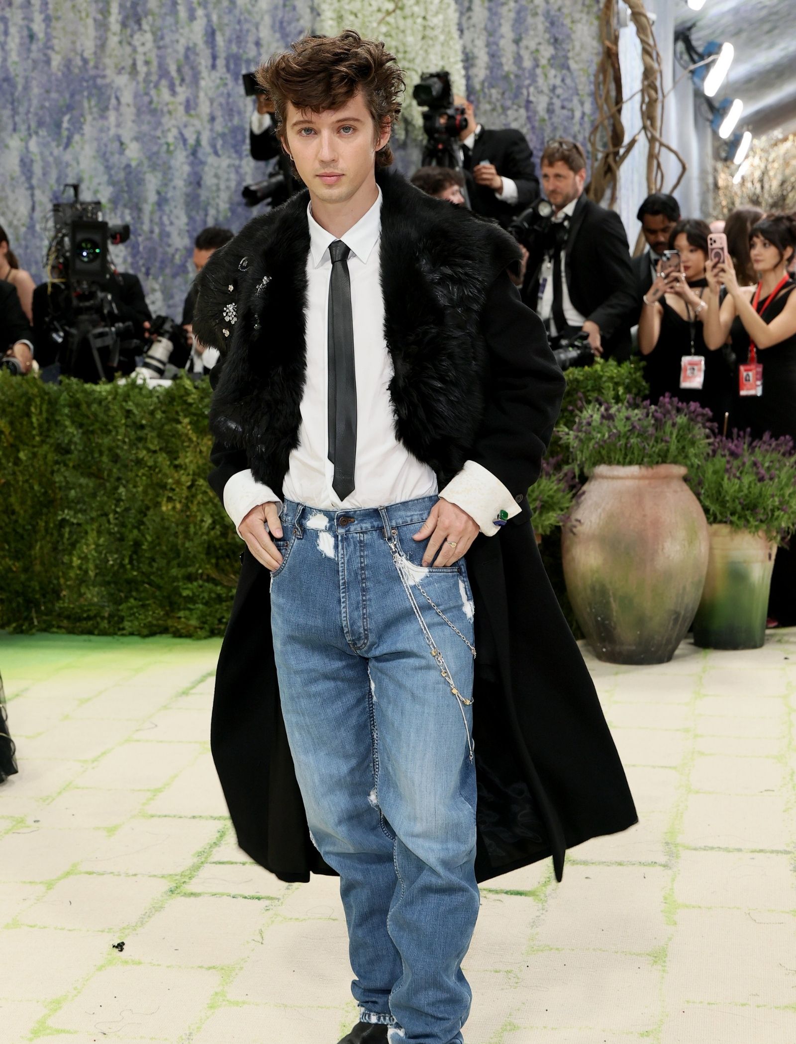 Male model walking on a runway dressed in a black fur coat, white shirt, black tie, and distressed jeans, with photographers in the background.