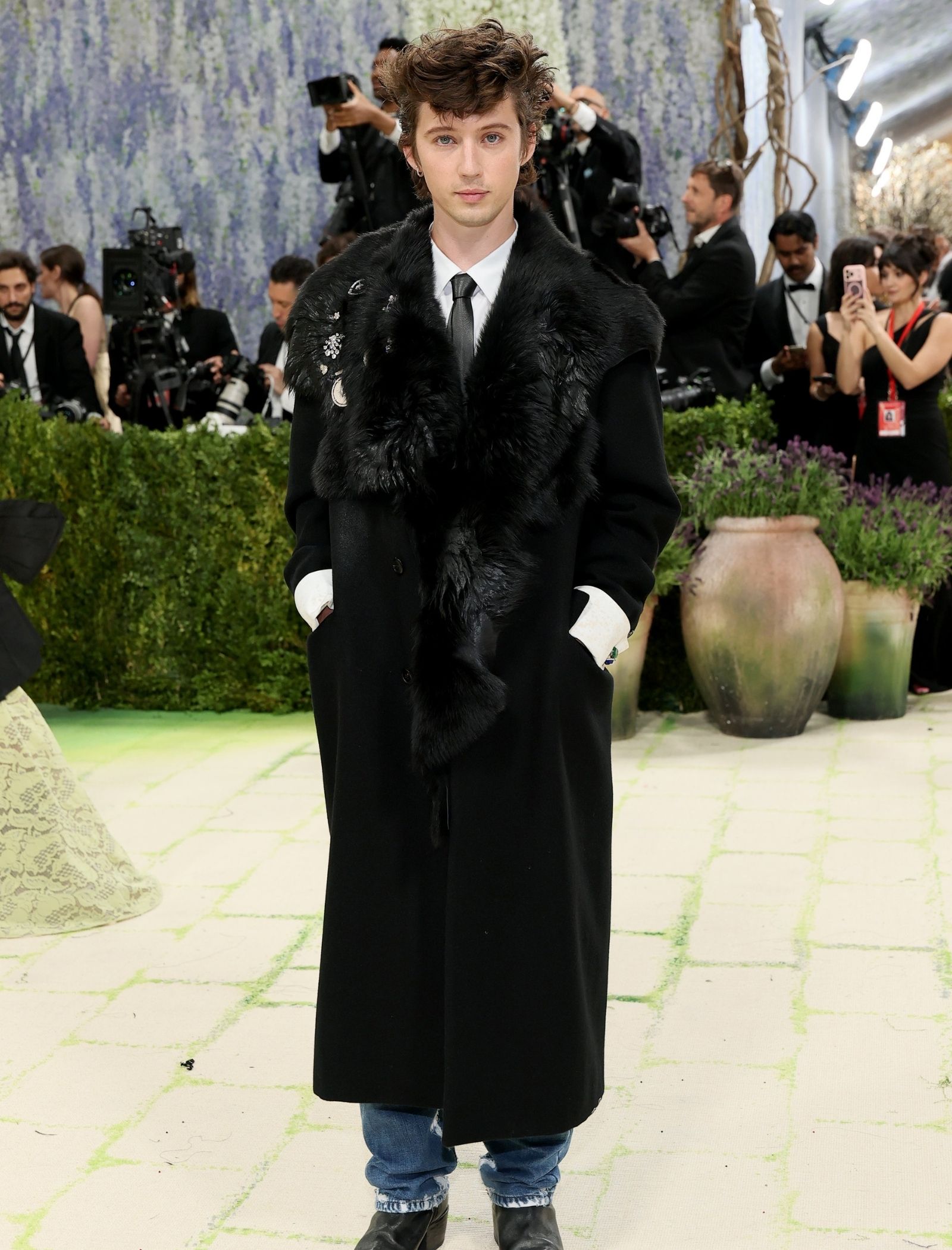 Person wearing a long black fur coat at a formal event, posing for photographers with a crowd in the background.