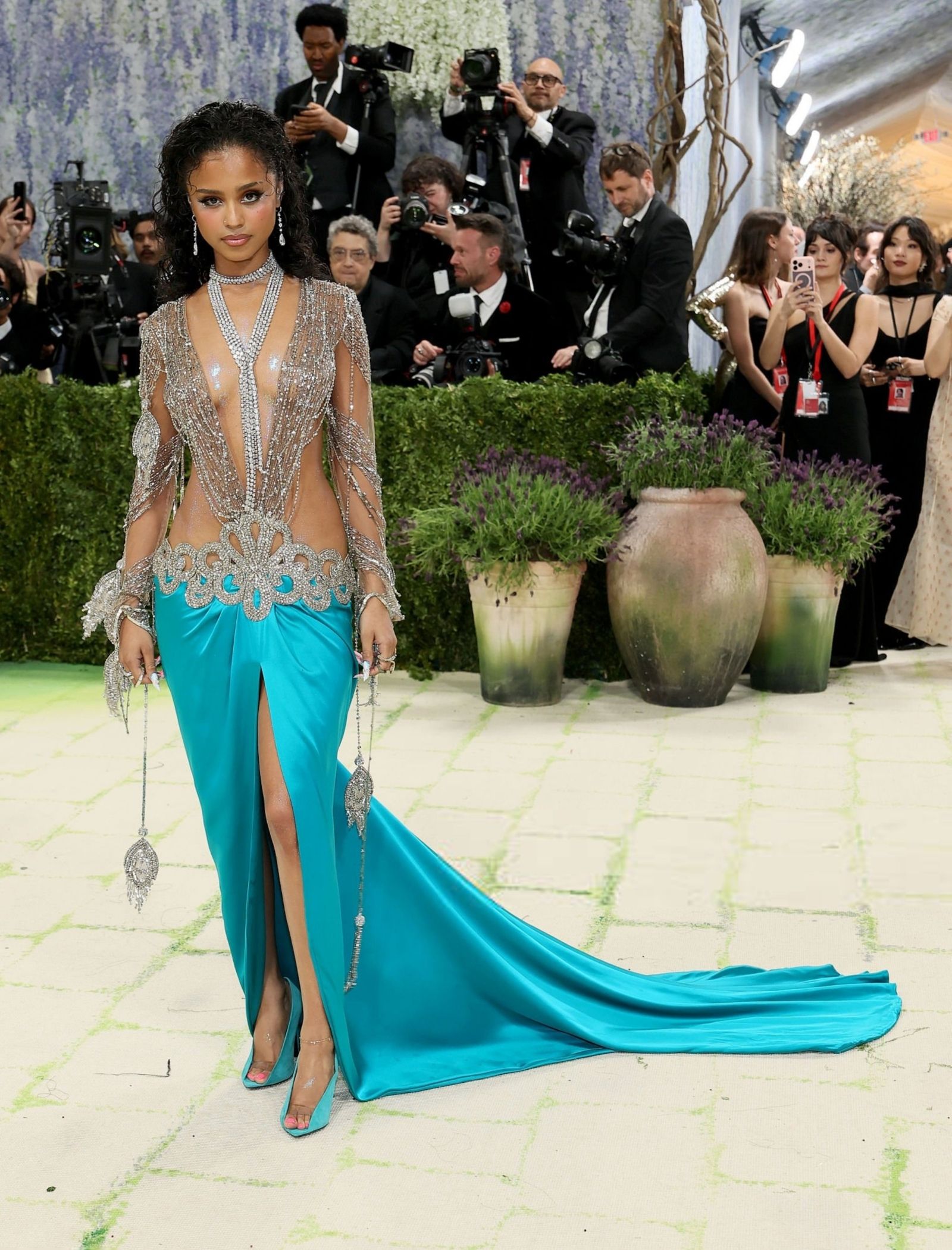 Model in a turquoise gown with a jeweled silver bodice walks the red carpet as photographers loom in the background.