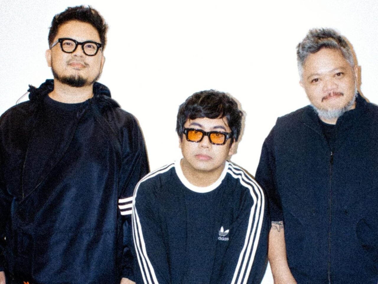 Three people posing together for a photo: two adults on the sides and a child in the middle, all wearing dark clothing and sunglasses (center kid with orange lenses).