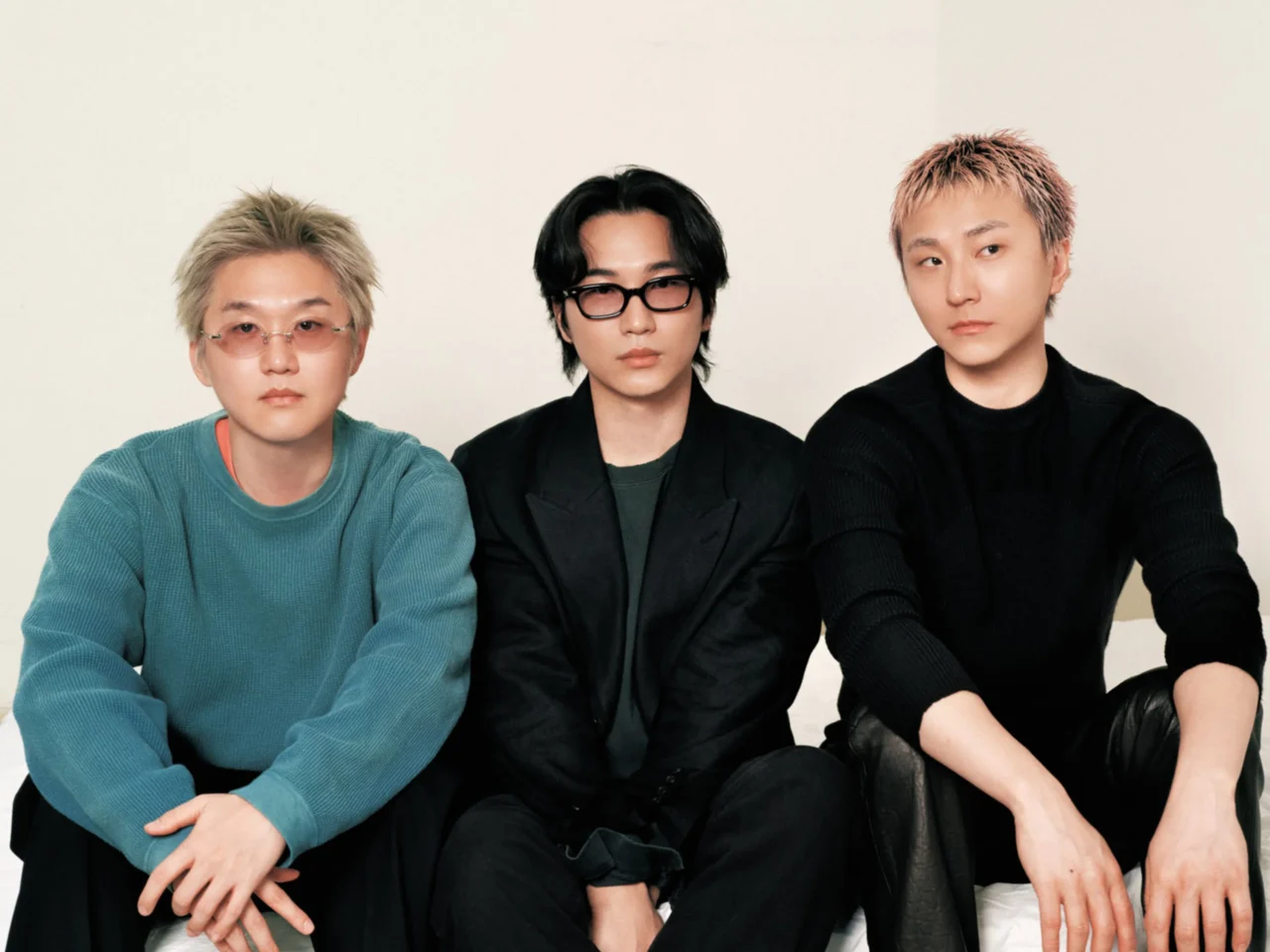 Three men sit together against a light background in casual outfits (left teal sweater, center black blazer, right black sweater).