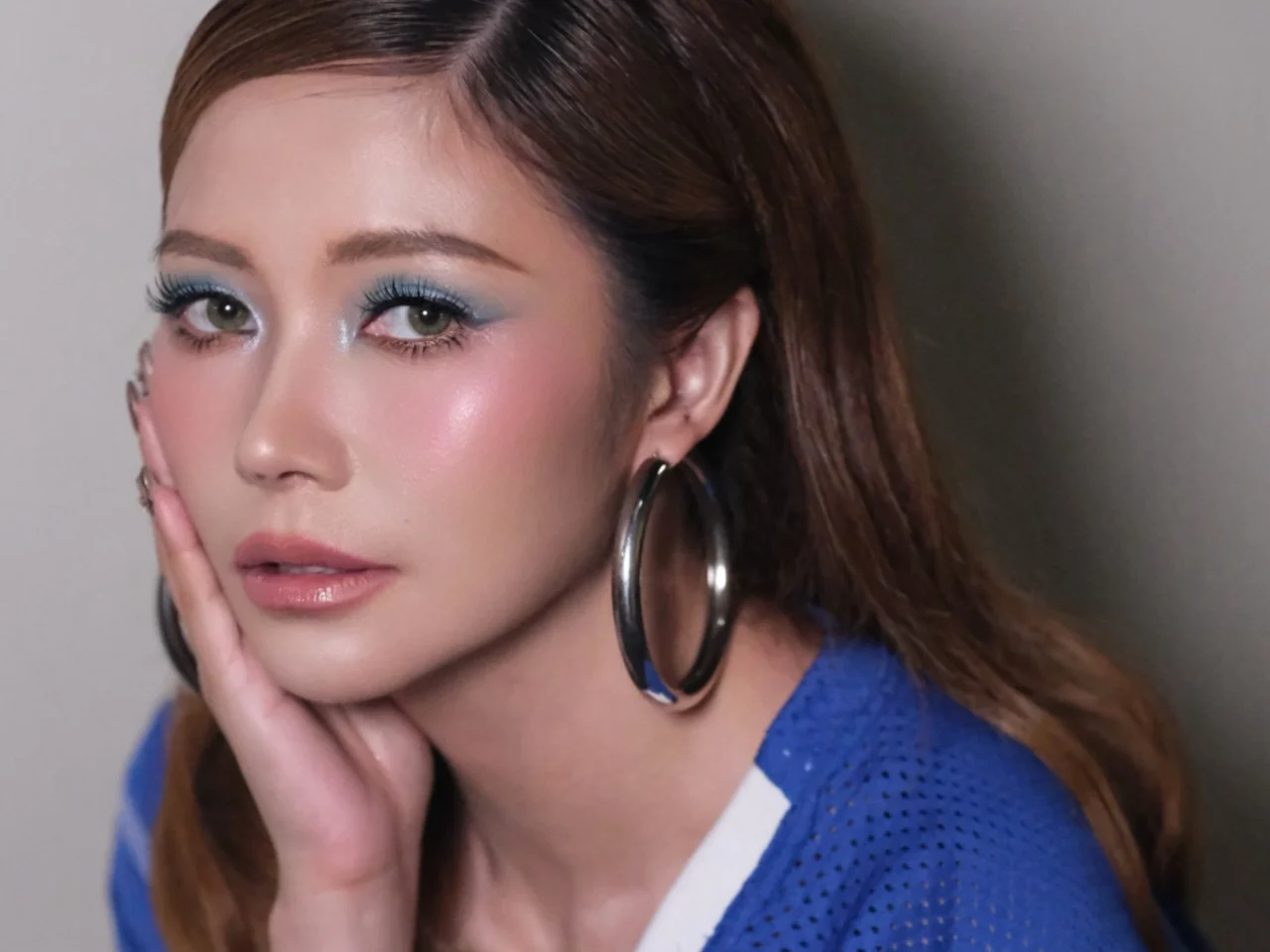 Close-up portrait of a woman with blue eyeshadow, glossy lips, and large hoop earrings, resting her cheek on her hand.