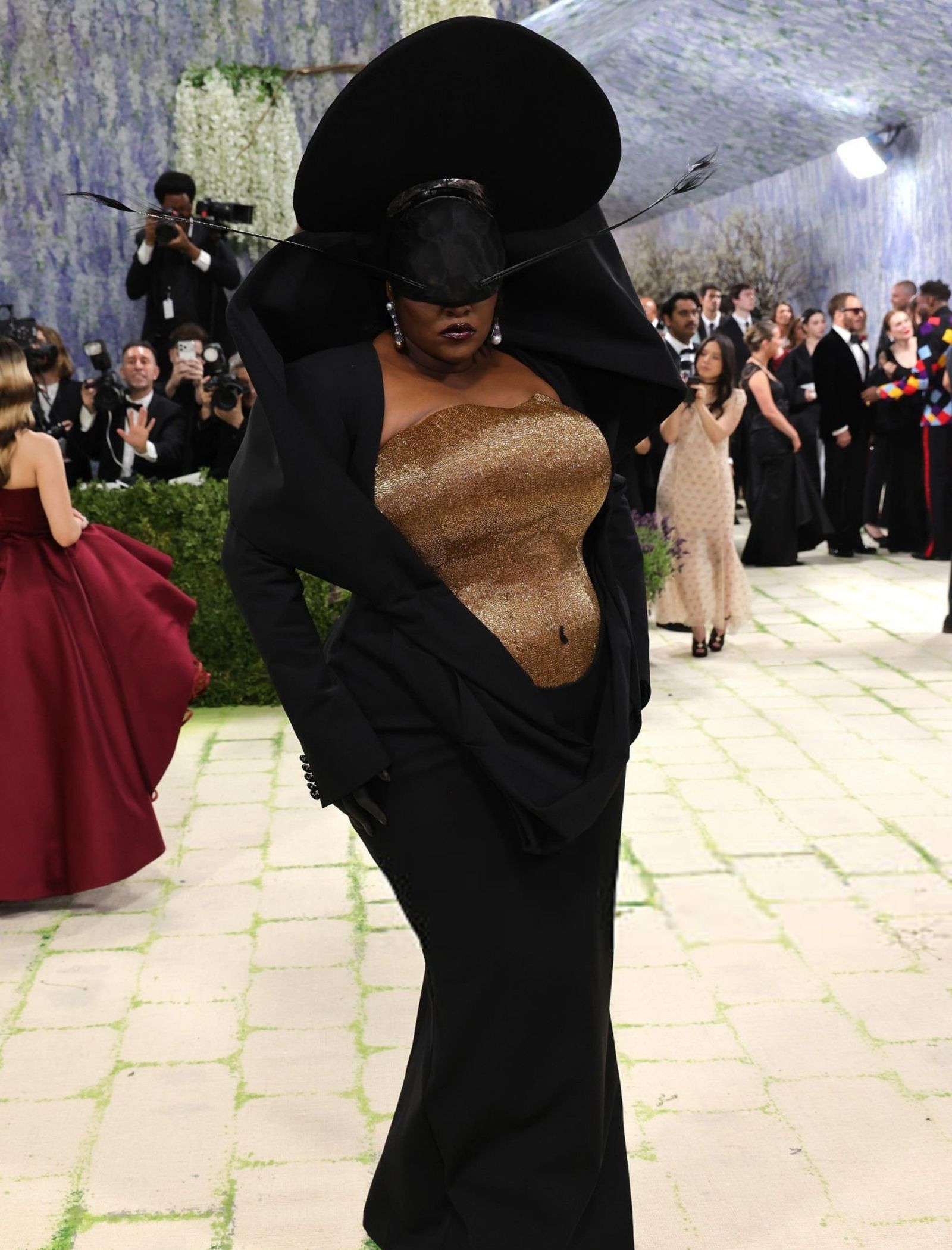 Model in a black gown with an oversized sculptural hat and gold glitter bodice on a fashion runway or red carpet, surrounded by photographers.