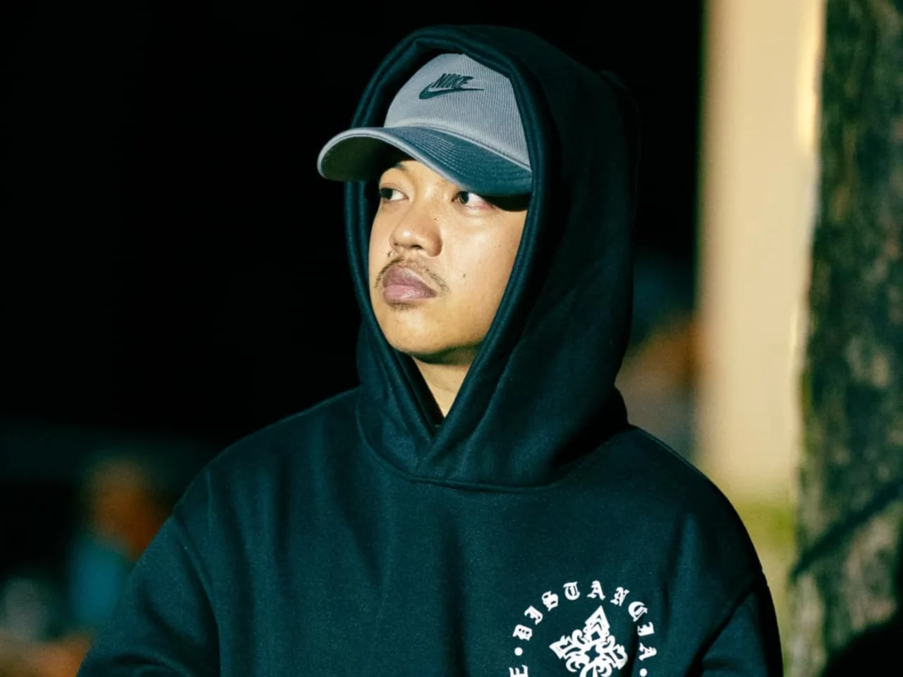 Young man wearing a black hoodie and gray Nike cap, looking to the left in a dark background.