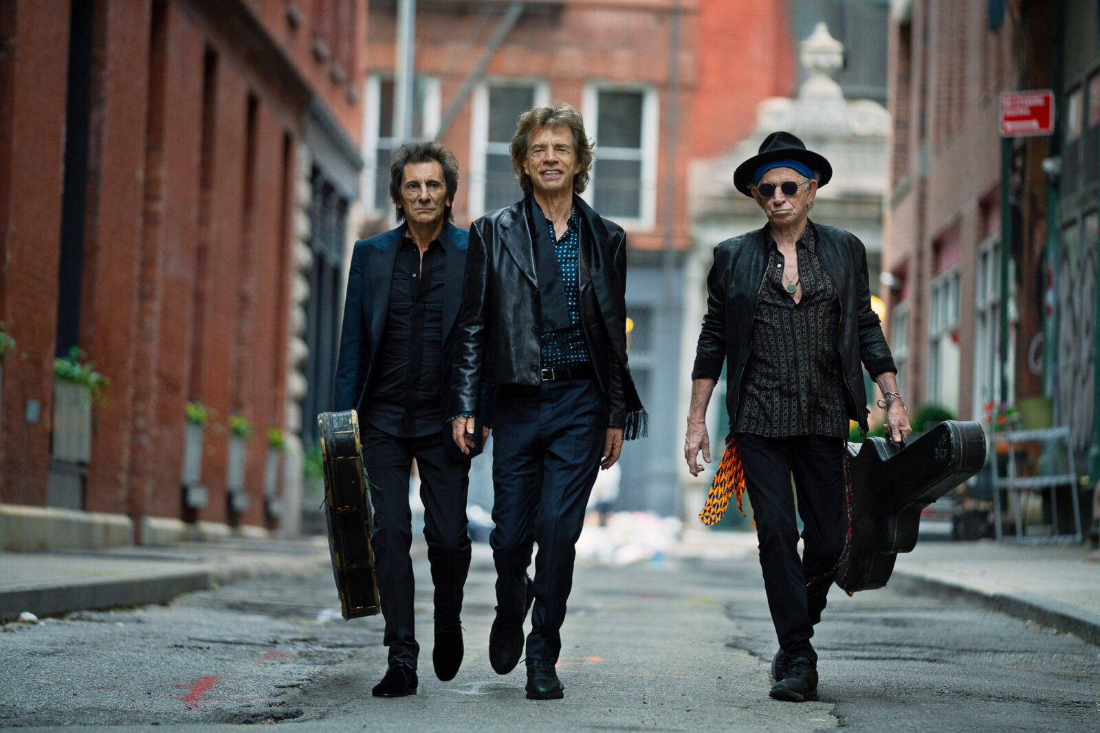 Ronnie Wood, Mick Jagger, and Keith Richards of the Rolling Stones walking on a street carrying guitar cases