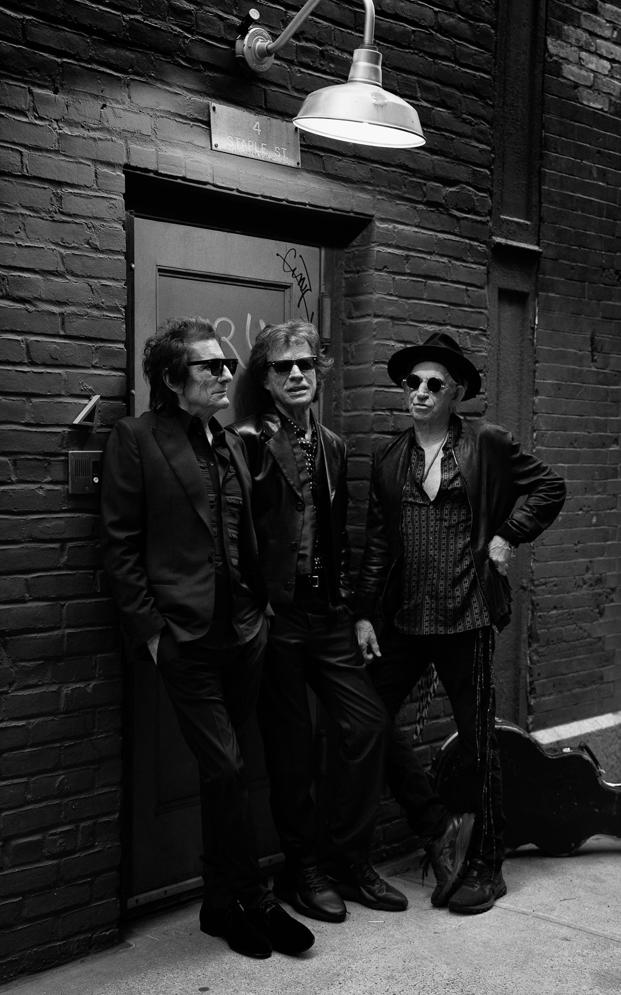 Black and white photo of Ronnie Wood, Mick Jagger, and Keith Richards of the Rolling Stones standing in front of a door on a brick wall